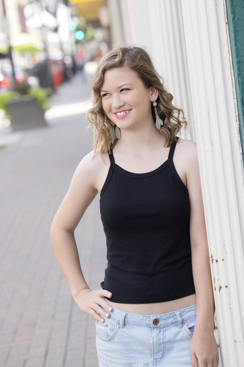 High school senior smiles confidently in a black tank top and light jeans while leaning against a downtown La Crosse building, captured by Mueller Photography