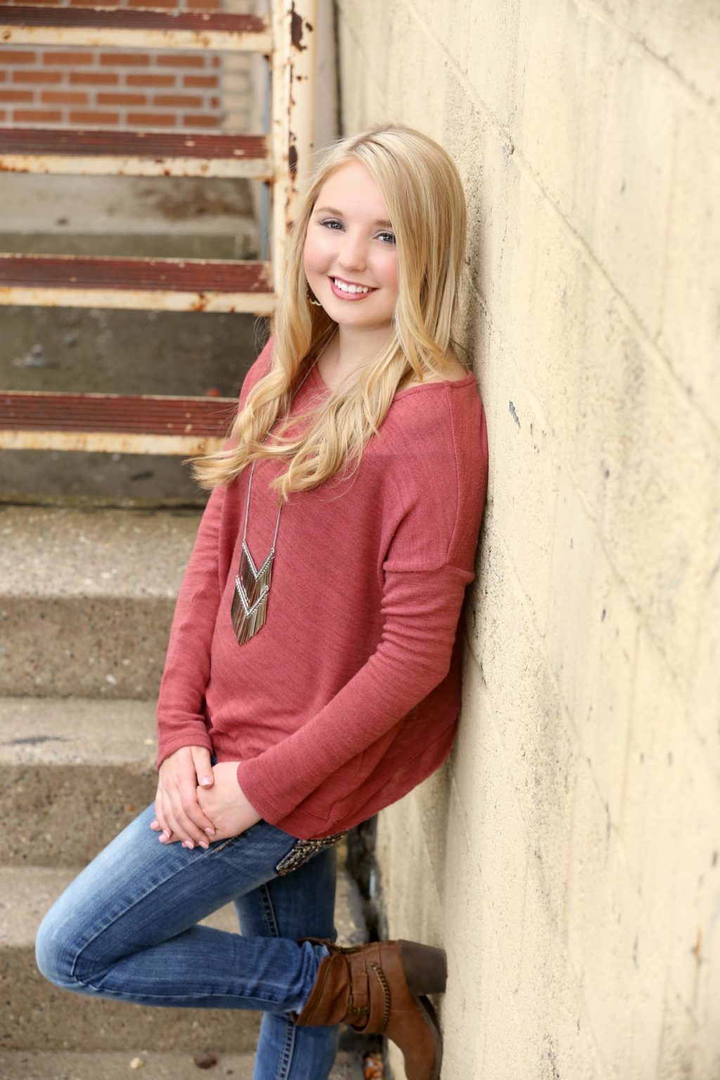 High school senior leans casually against a concrete wall near an old stairway, smiling during an urban senior portrait session with Mueller Photography in La Crosse, WI.