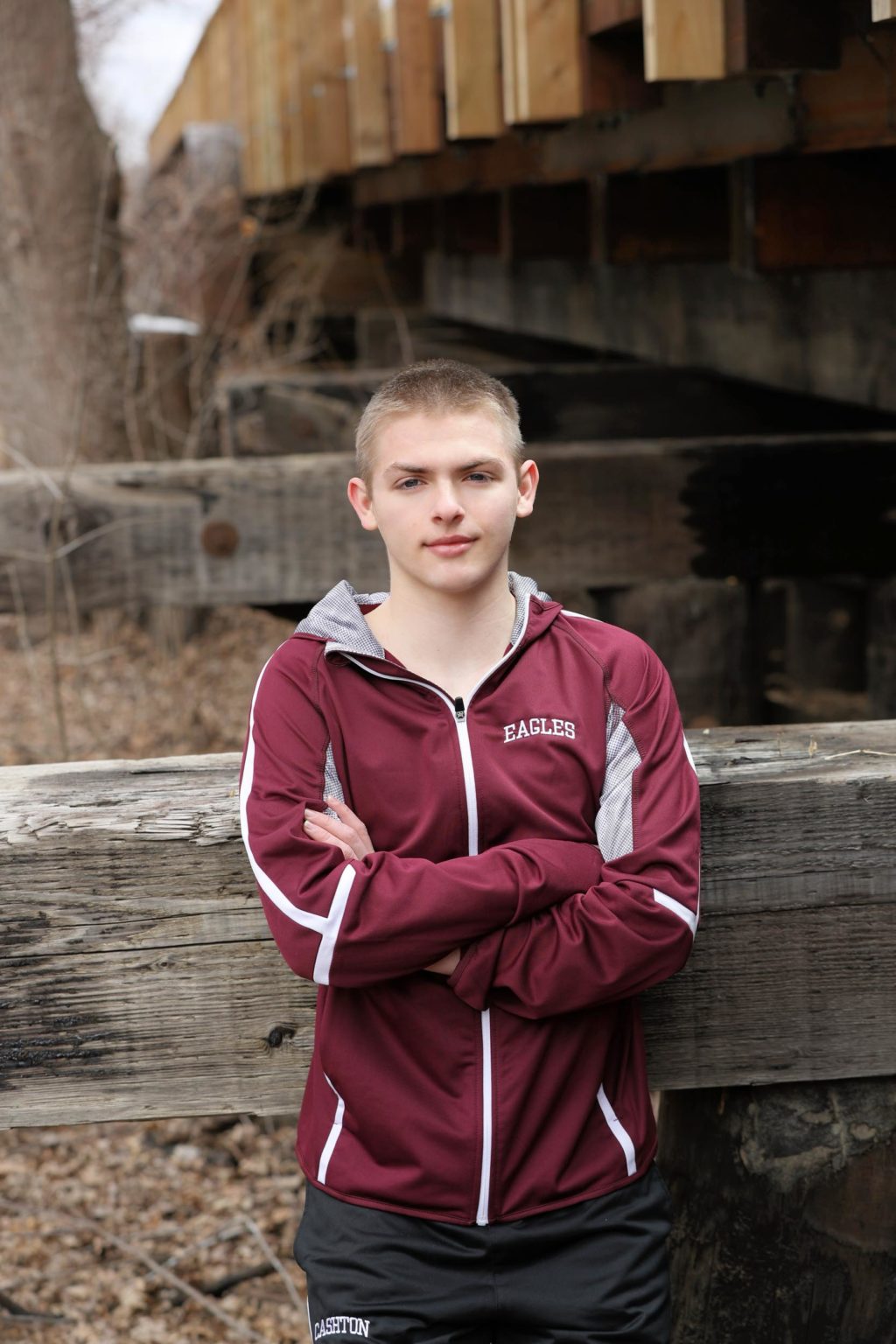 High school senior stands with arms crossed in a maroon athletic jacket under a wooden bridge during a cool-season senior portrait session with Mueller Photography in La Crosse, WI.