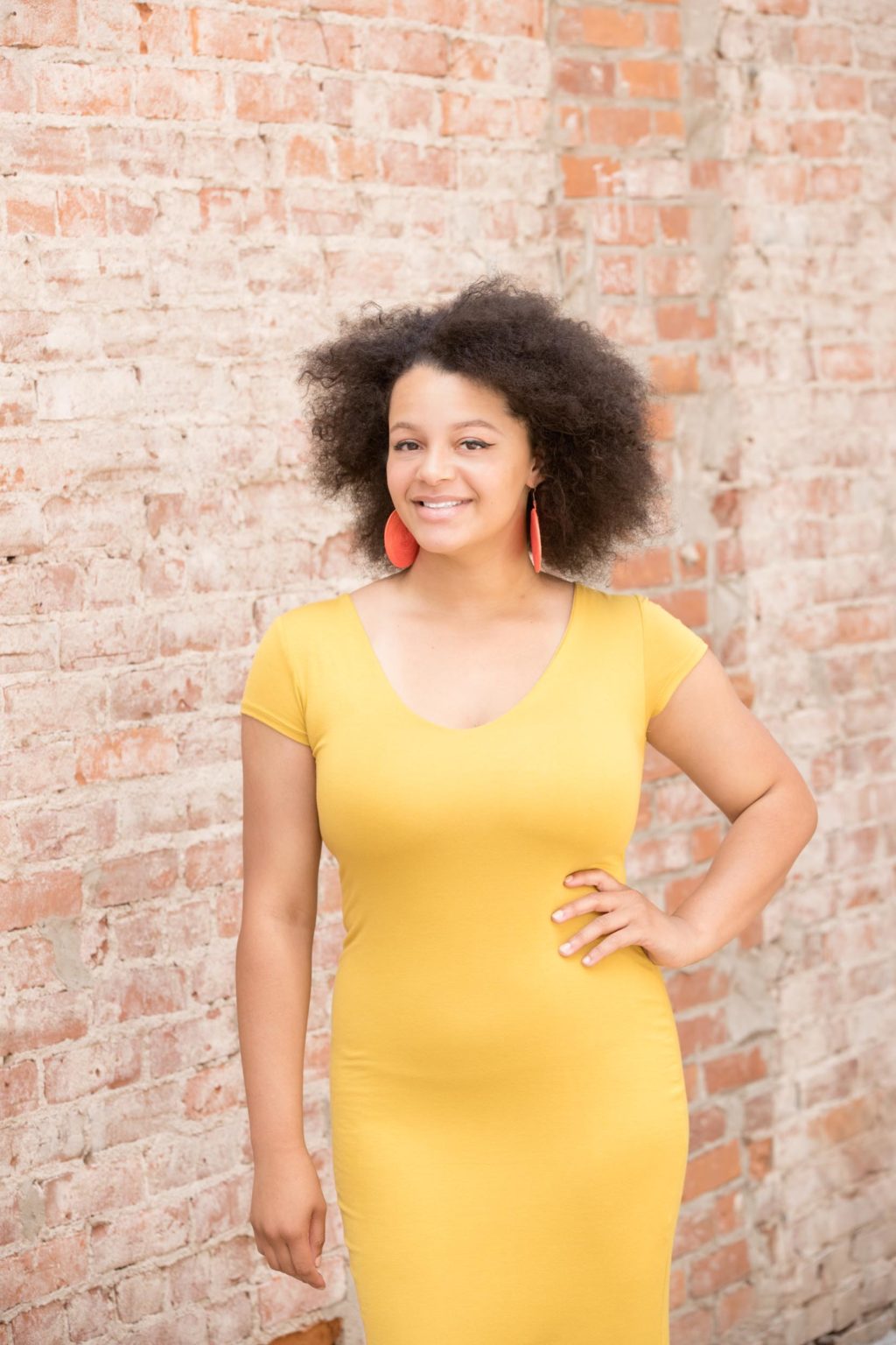 High school senior smiles confidently in a bold yellow dress with red earrings during an urban senior portrait session with Mueller Photography in La Crosse, WI.