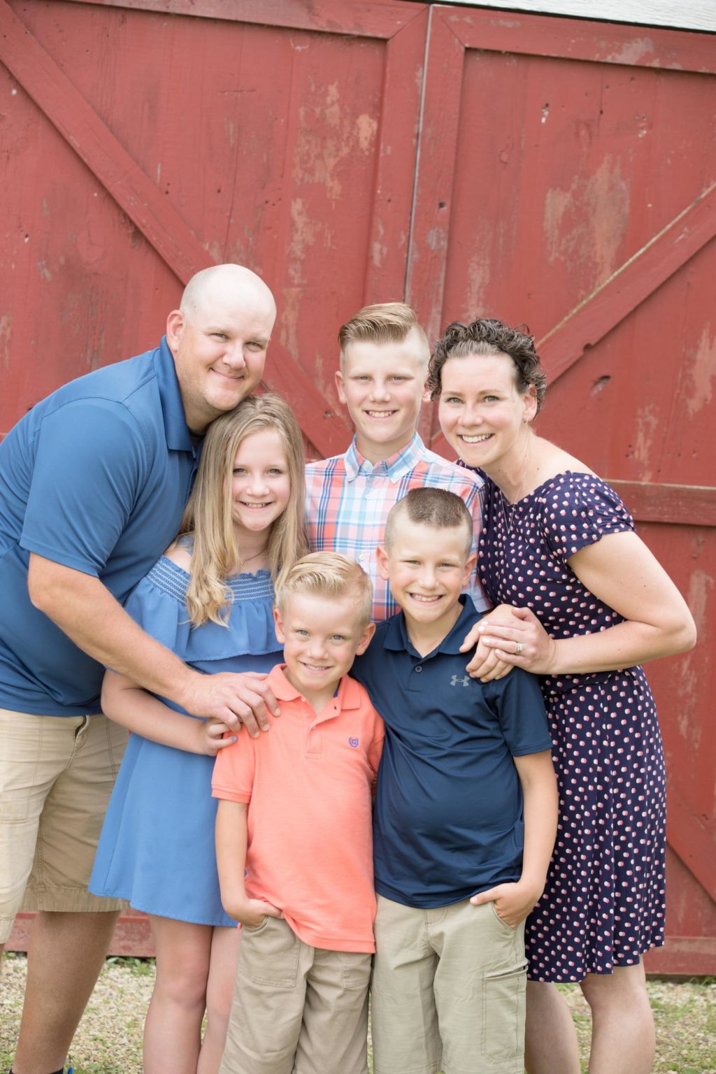 Family of six smiles together in front of a rustic red barn during a summer portrait session with Mueller Photography in La Crosse, WI.