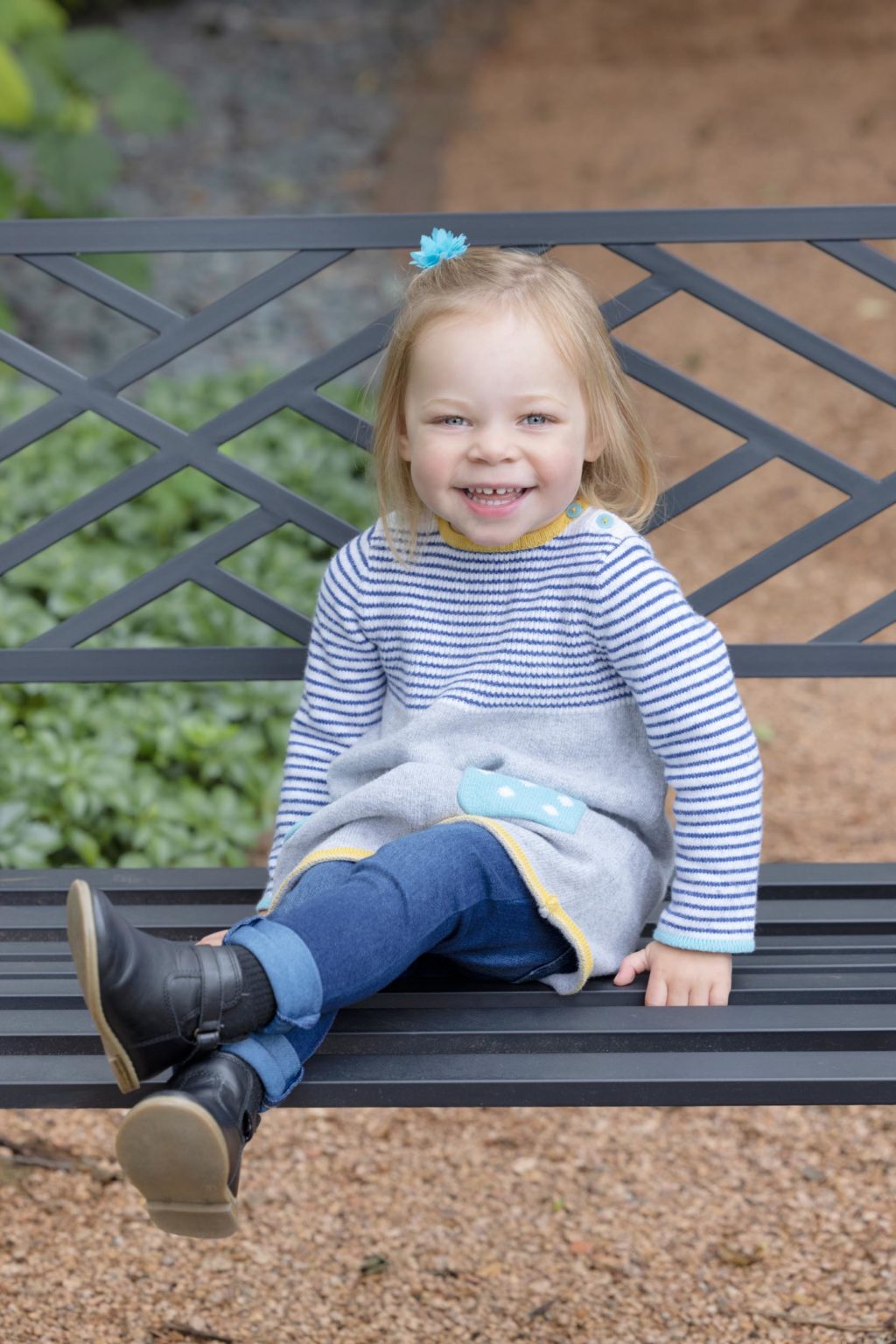 Young girl smiling while sitting on a bench during outdoor family portraits in La Crosse WI, dressed in a striped sweater and jeans with boots