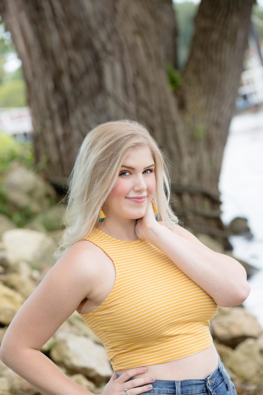 High school senior poses confidently in a yellow top and jeans near a riverside backdrop with large trees during a portrait session with Mueller Photography in La Crosse, WI.