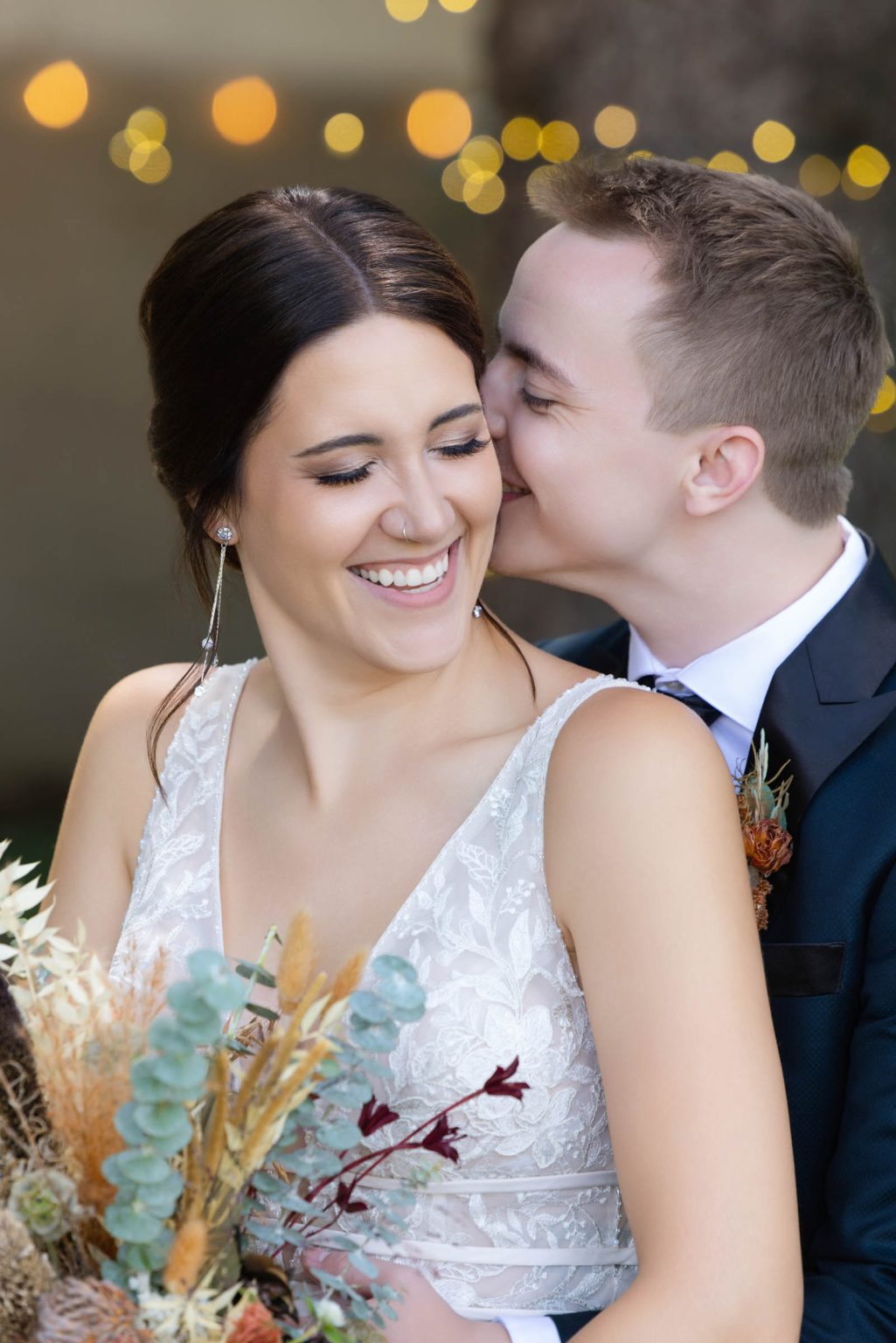 Bride laughs as groom kisses her cheek during golden hour portraits in Madison, WI, holding a fall-inspired bouquet, captured by Mueller Photography