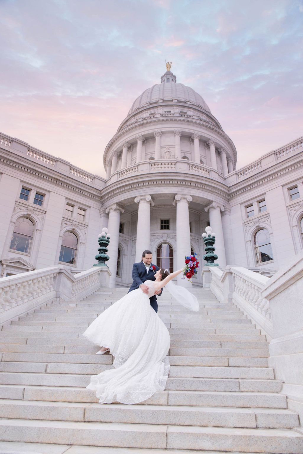 Groom dips bride on the grand steps of the Wisconsin State Capitol at sunset, her gown flowing dramatically, captured by Mueller Photography