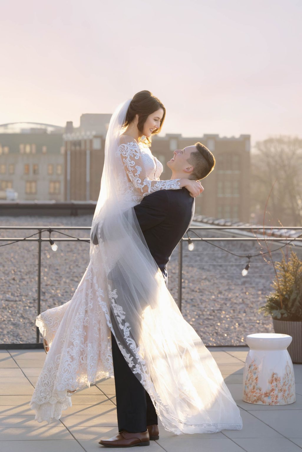 Groom lifts bride in a joyful embrace on a rooftop at sunset, her lace gown flowing as they smile at each other, captured by Mueller Photography in La Crosse WI