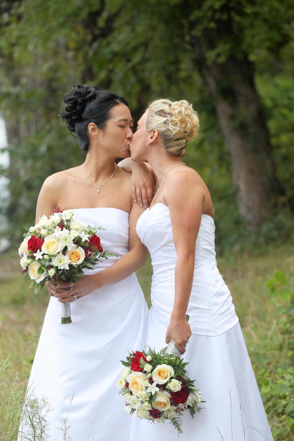 Two brides share a kiss while holding floral bouquets in a natural setting at Pettibone Park in La Crosse, WI, captured by Mueller Photography