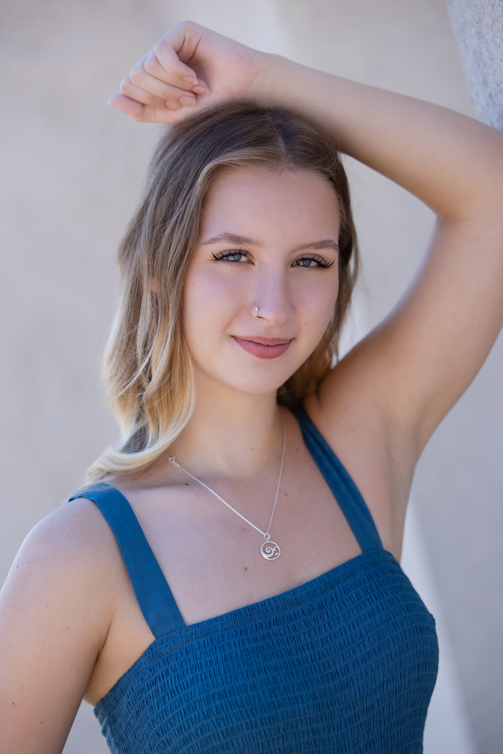 A young woman in a blue sleeveless dress poses with her arm resting on her head, looking directly at the camera with a soft smile.