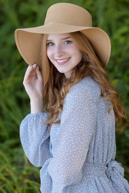High school senior smiles while wearing a wide-brimmed hat and soft blue dress during a summer senior portrait session with Mueller Photography in La Crosse, WI.
