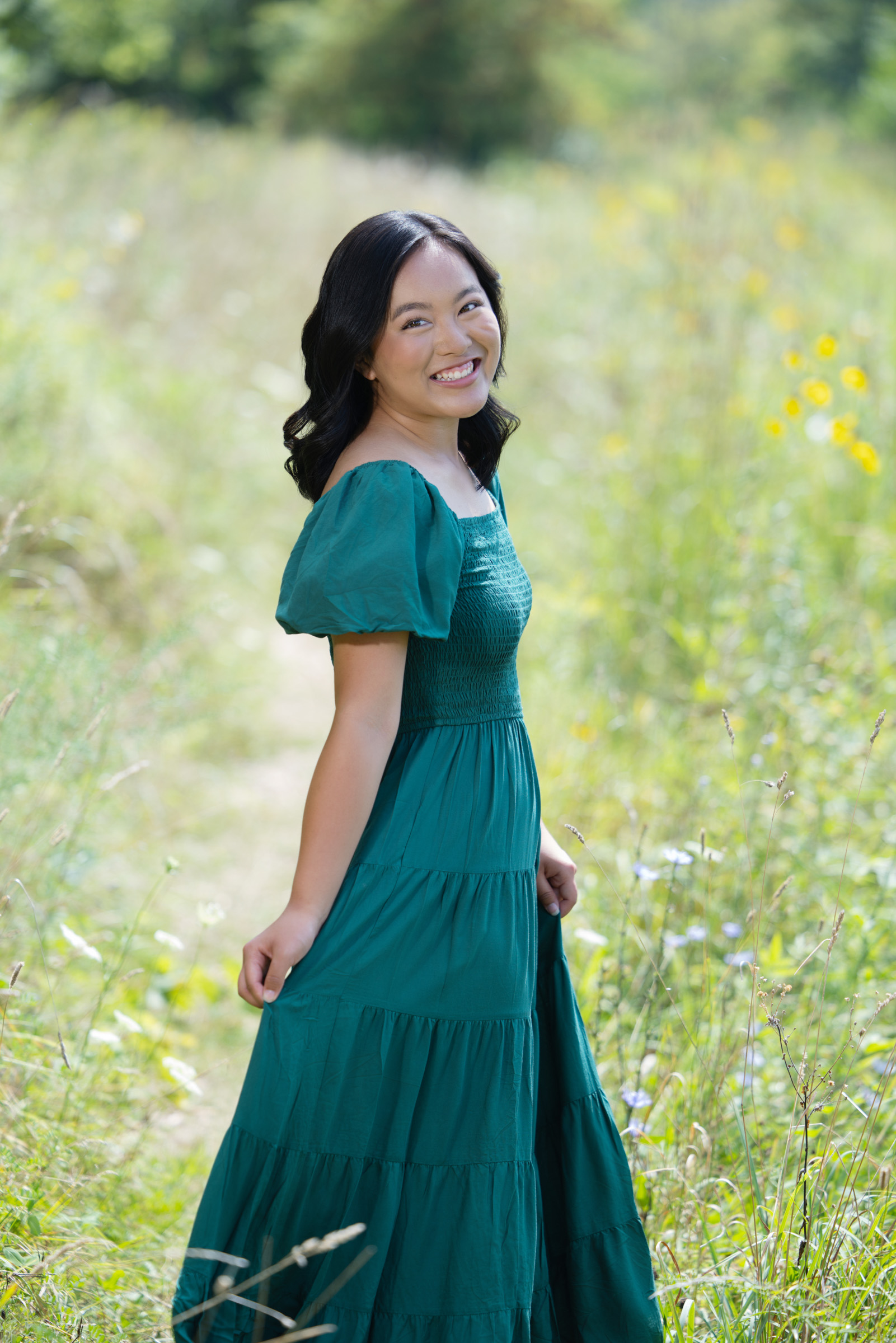 High school senior smiles in a flowing emerald green dress, standing along a wildflower-lined path during a summer portrait session with Mueller Photography in La Crosse, WI.