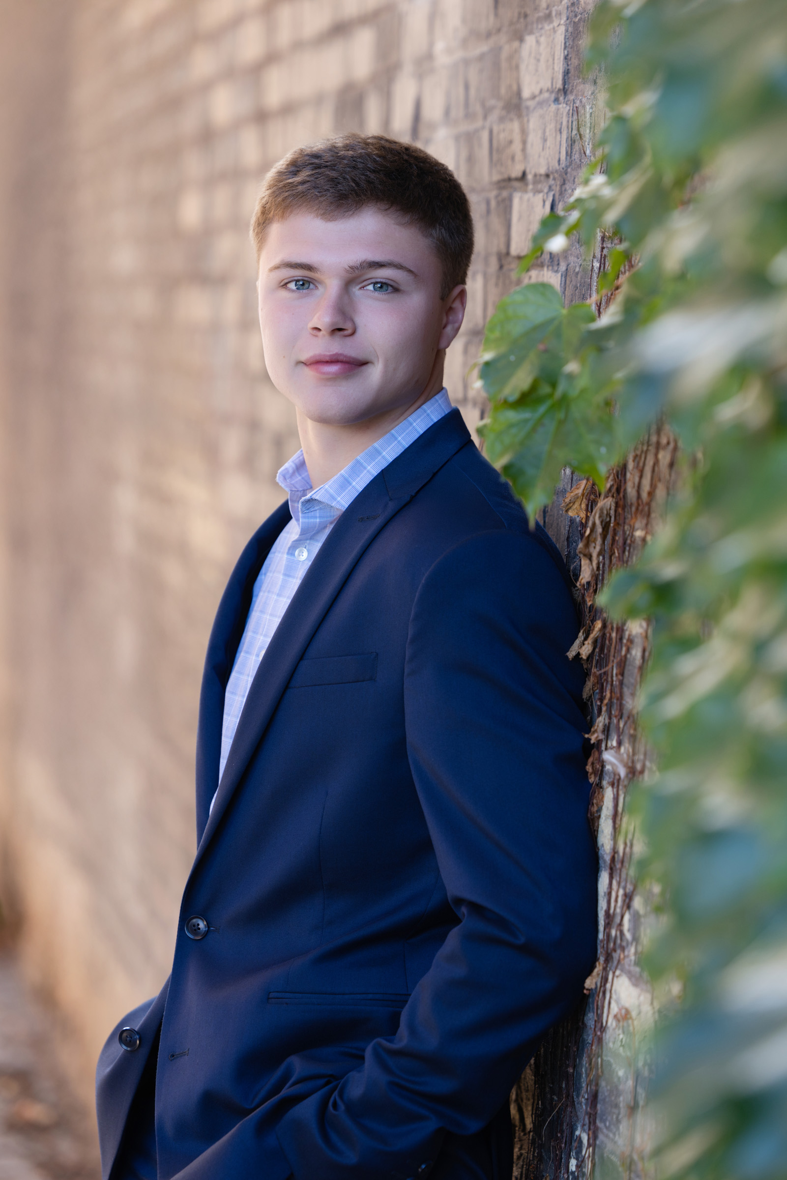High school senior dressed in a navy blazer poses confidently against a brick wall with ivy during a fall portrait session with Mueller Photography in La Crosse, WI.