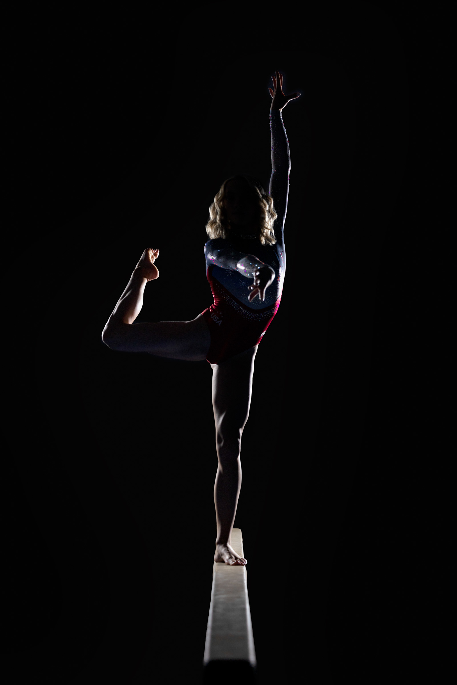Gymnast poses in a dramatic spotlight on the balance beam, perfectly centered in a graceful arch during a gymnastics portrait session in Minneapolis with Mueller Photography.