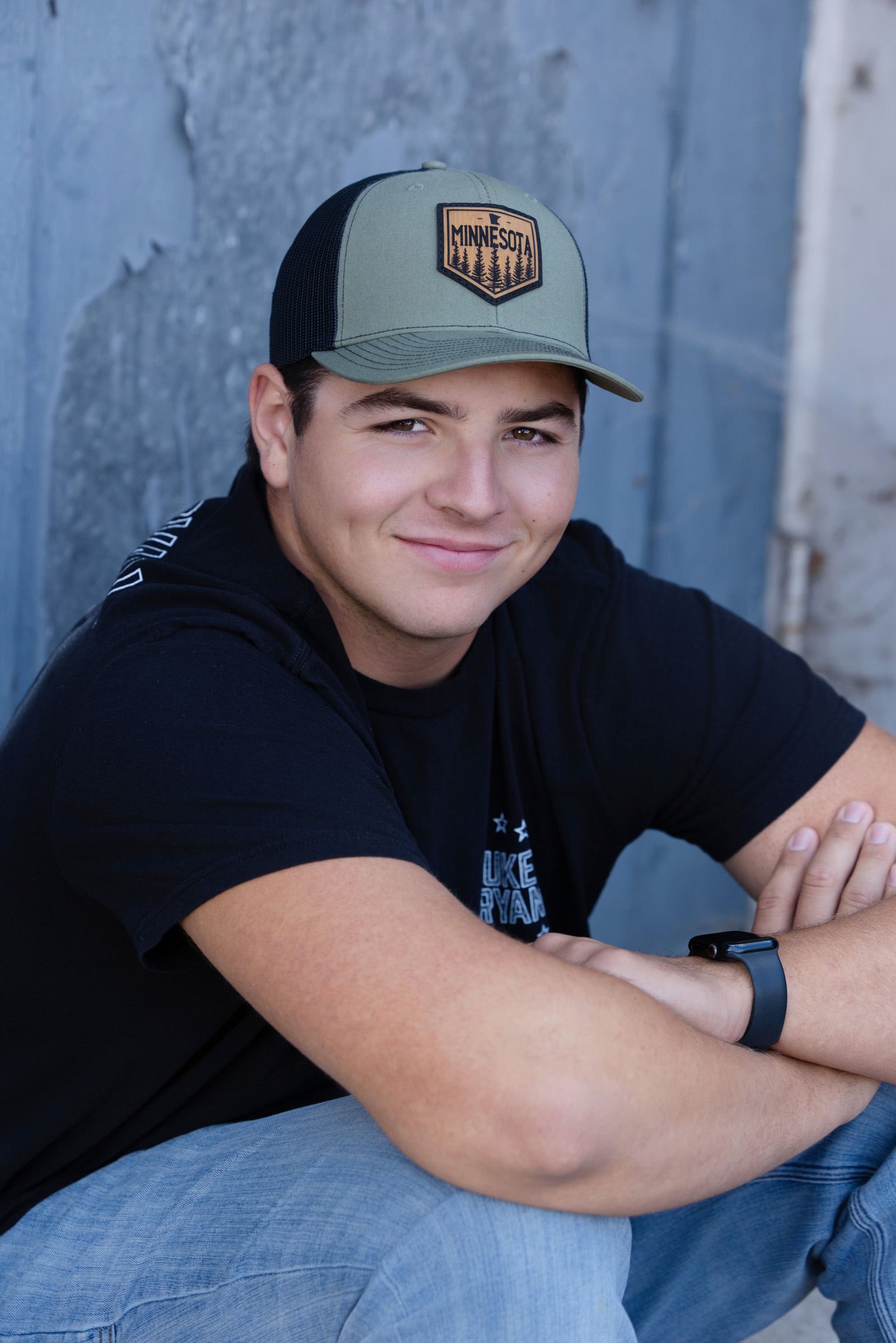 Outdoor senior portrait of a young man in a casual black shirt and cap, leaning against a blue wall with a relaxed smile during his La Crosse WI session.