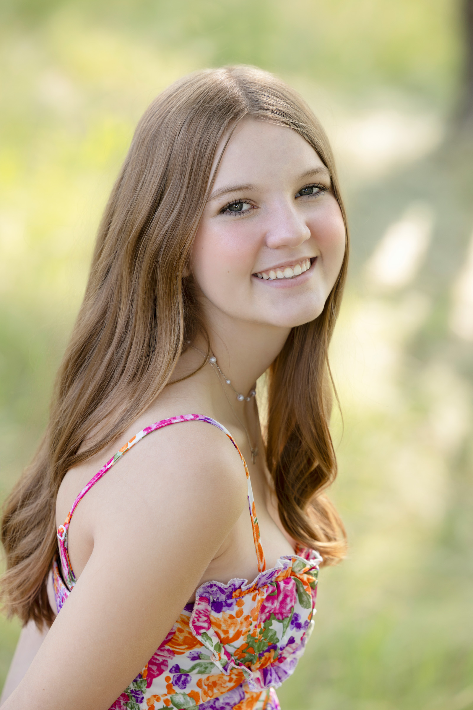High school senior with long hair smiles softly in a floral dress, surrounded by soft greenery and warm summer light during her senior portrait session in La Crosse, WI.