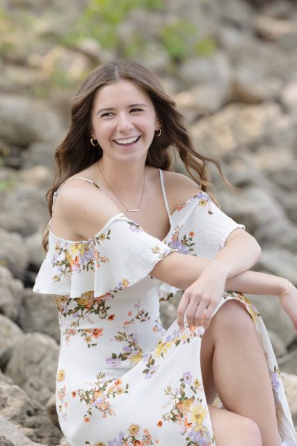 High school senior smiles while sitting on a rocky shoreline in a floral dress during a natural light senior portrait session with Mueller Photography in La Crosse, WI.