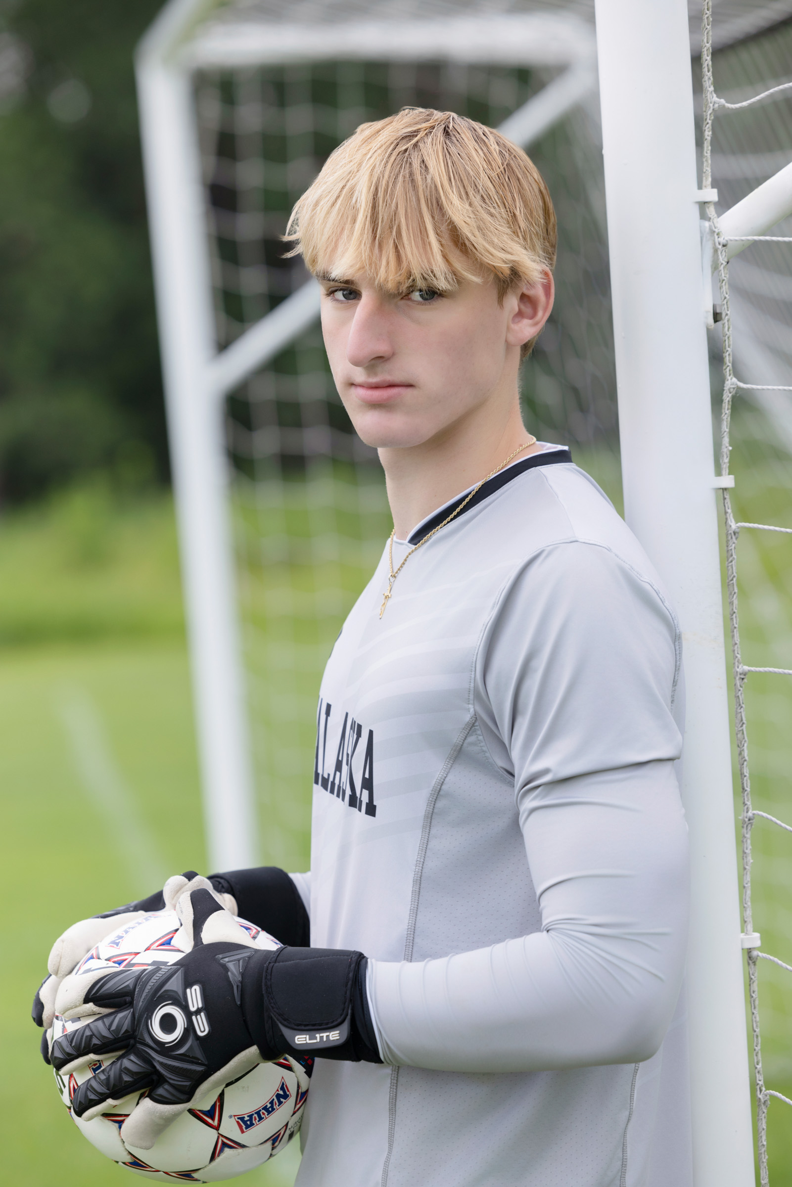 High school senior in goalkeeper uniform stands confidently at the goalpost holding a soccer ball, captured during a sports-themed senior portrait session in La Crosse, WI.