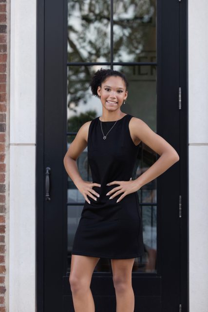 High school senior stands confidently in a black dress during a downtown portrait session with Mueller Photography in La Crosse, WI, posing in front of elegant black doors.