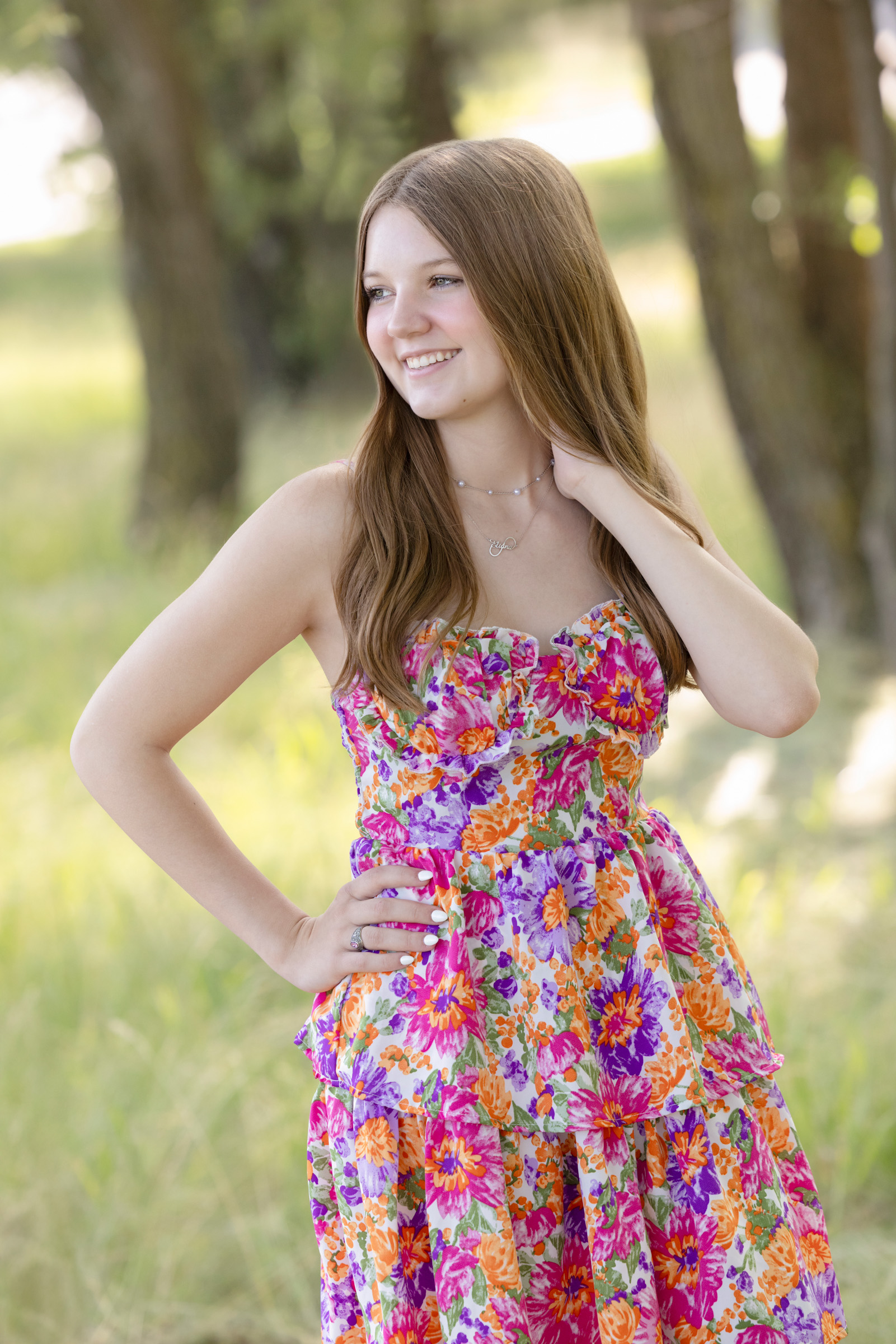 Senior girl in a colorful floral dress stands in a sunlit grassy field, smiling off-camera with one hand on her hip during a summer portrait session in La Crosse, WI.