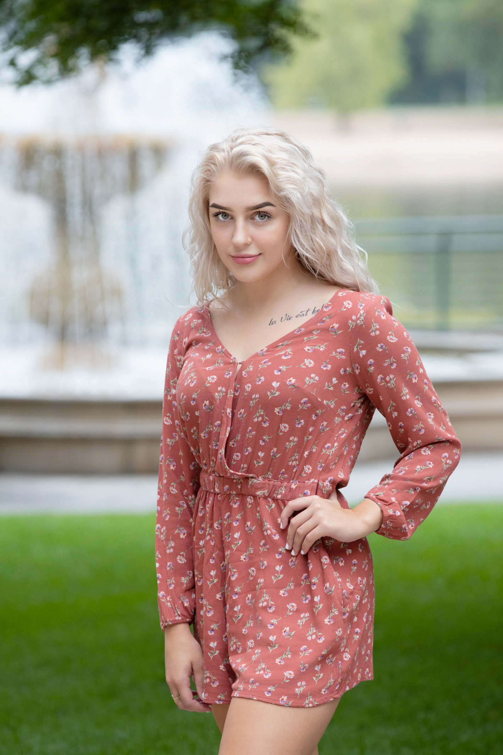 Senior girl with platinum blonde hair poses confidently in a rust-colored floral romper with a fountain blurred in the background during her portrait session in La Crosse, WI.