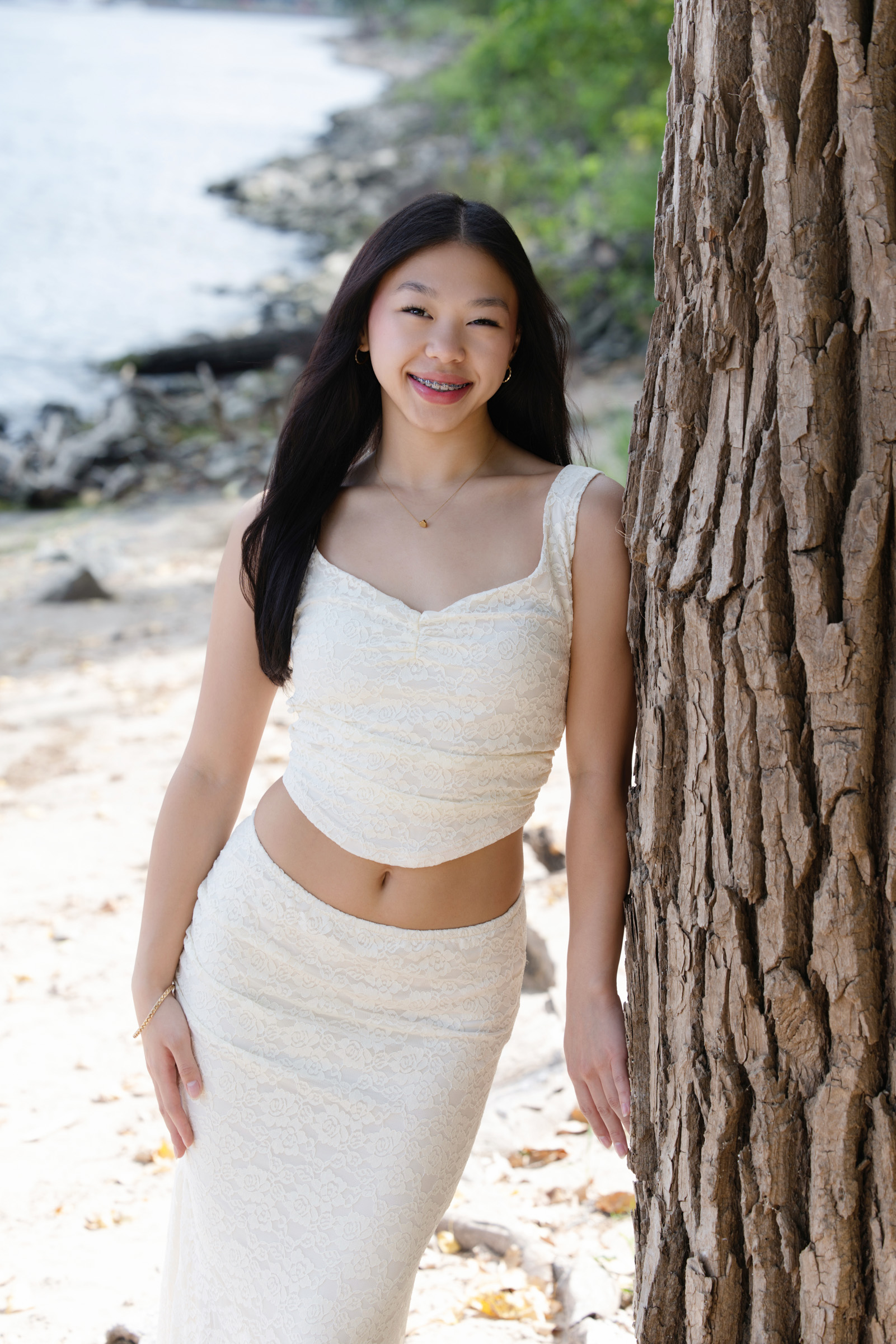 Senior girl in a white two-piece outfit leans against a tree on a sandy riverbank, smiling softly during her portrait session with Mueller Photography in La Crosse, WI.