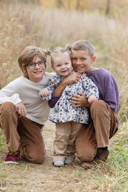 Two older children sitting with their younger sister during an outdoor family portrait.