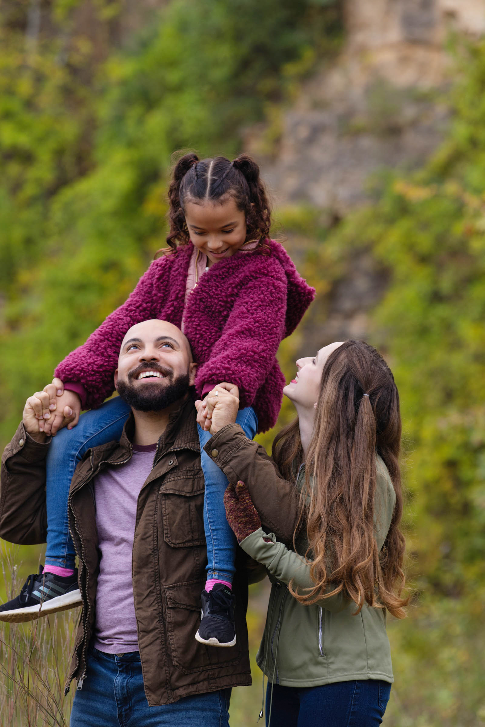 Parents smiling up at their daughter as she sits on her father's shoulders during an outdoor family portrait session.