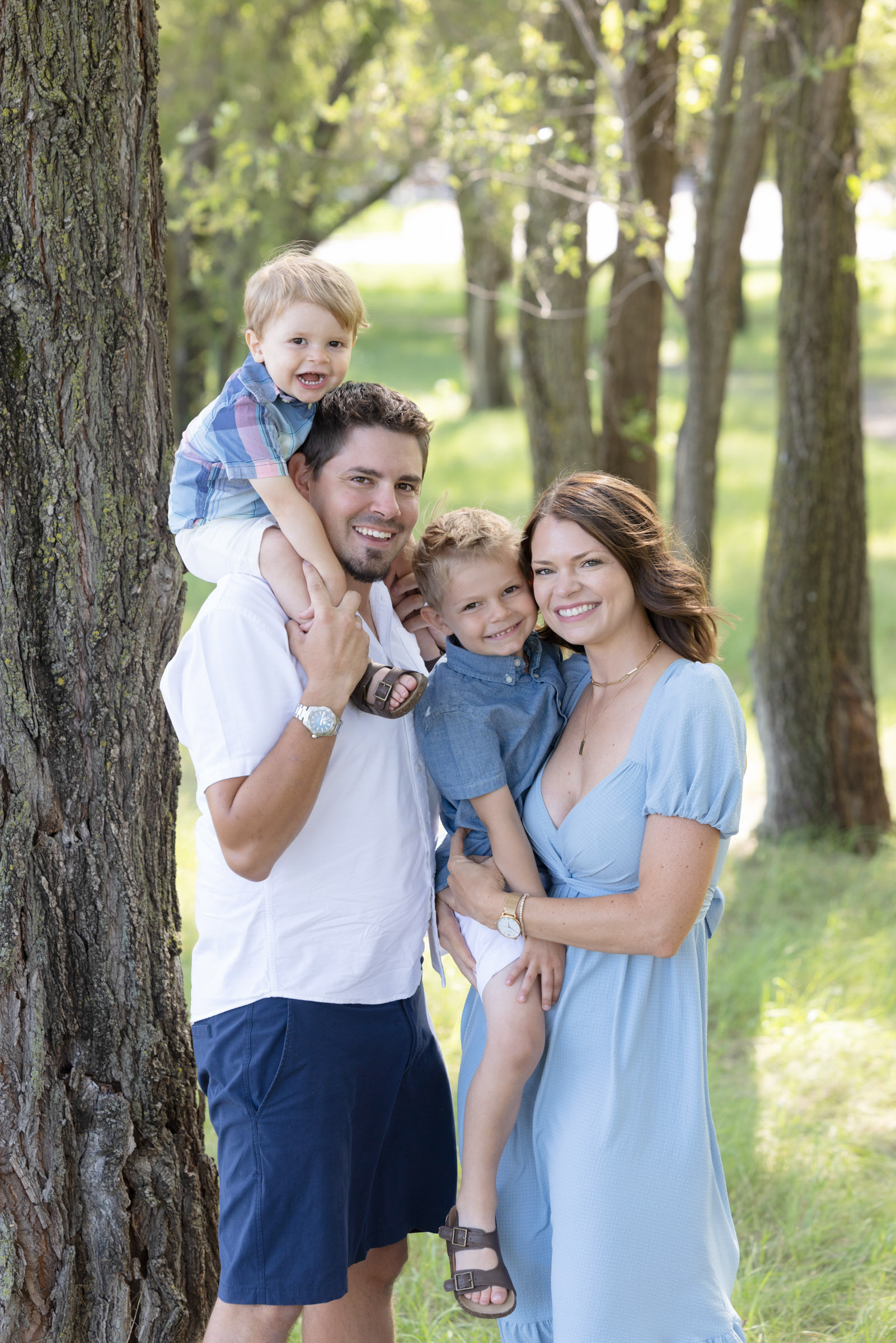 Parents carrying their children while walking together through a wooded park during a family portrait session.