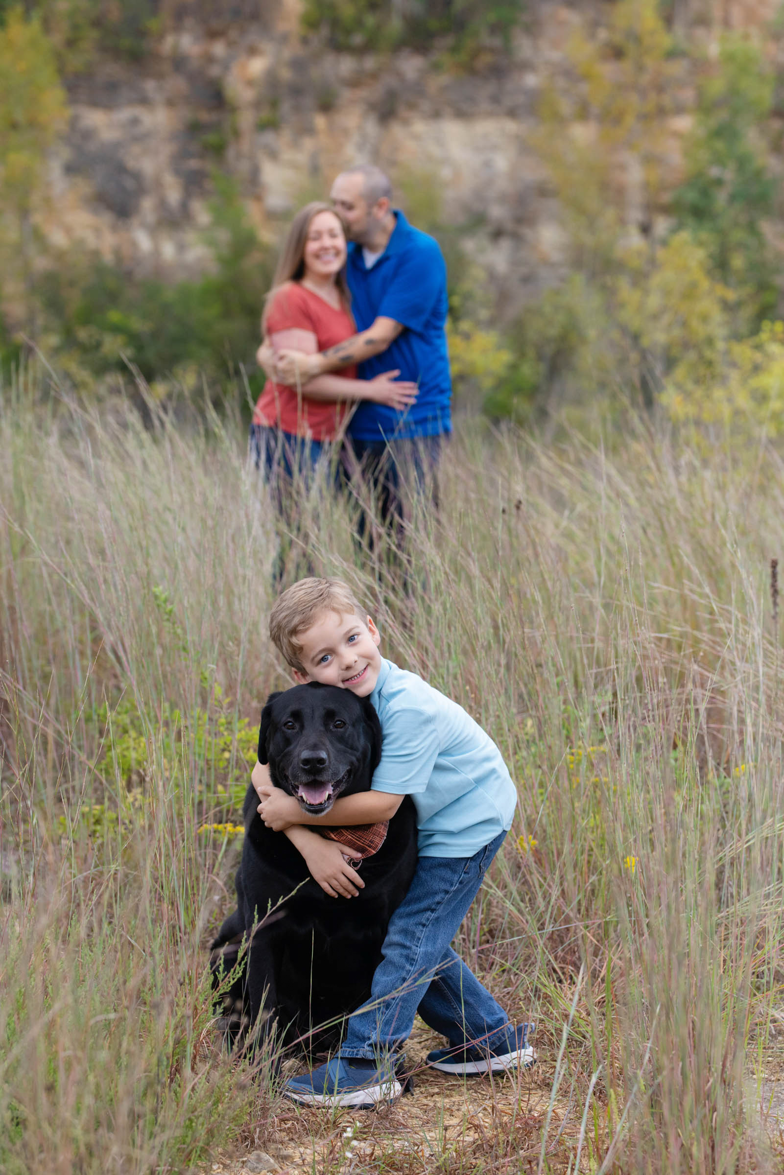 Parents standing behind their child, who is hugging the family dog in a grassy field during an outdoor portrait session.
