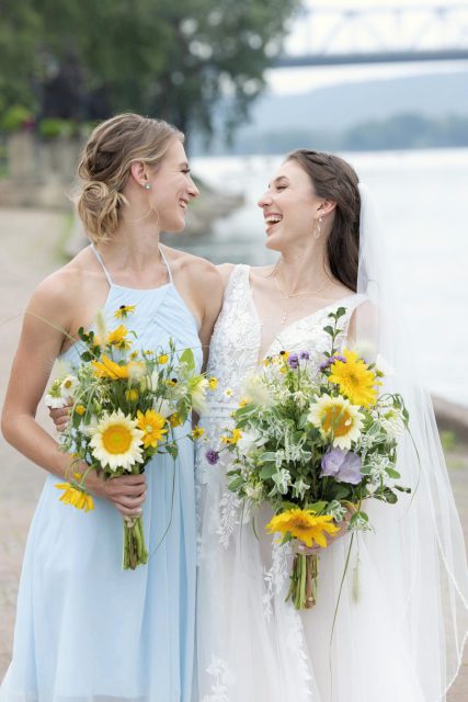 Bride and bridesmaid smiling together while holding bouquets before the ceremony.