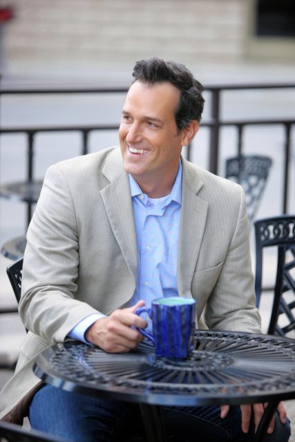 Professional portrait of a man seated outdoors with a coffee mug, smiling toward the camera.