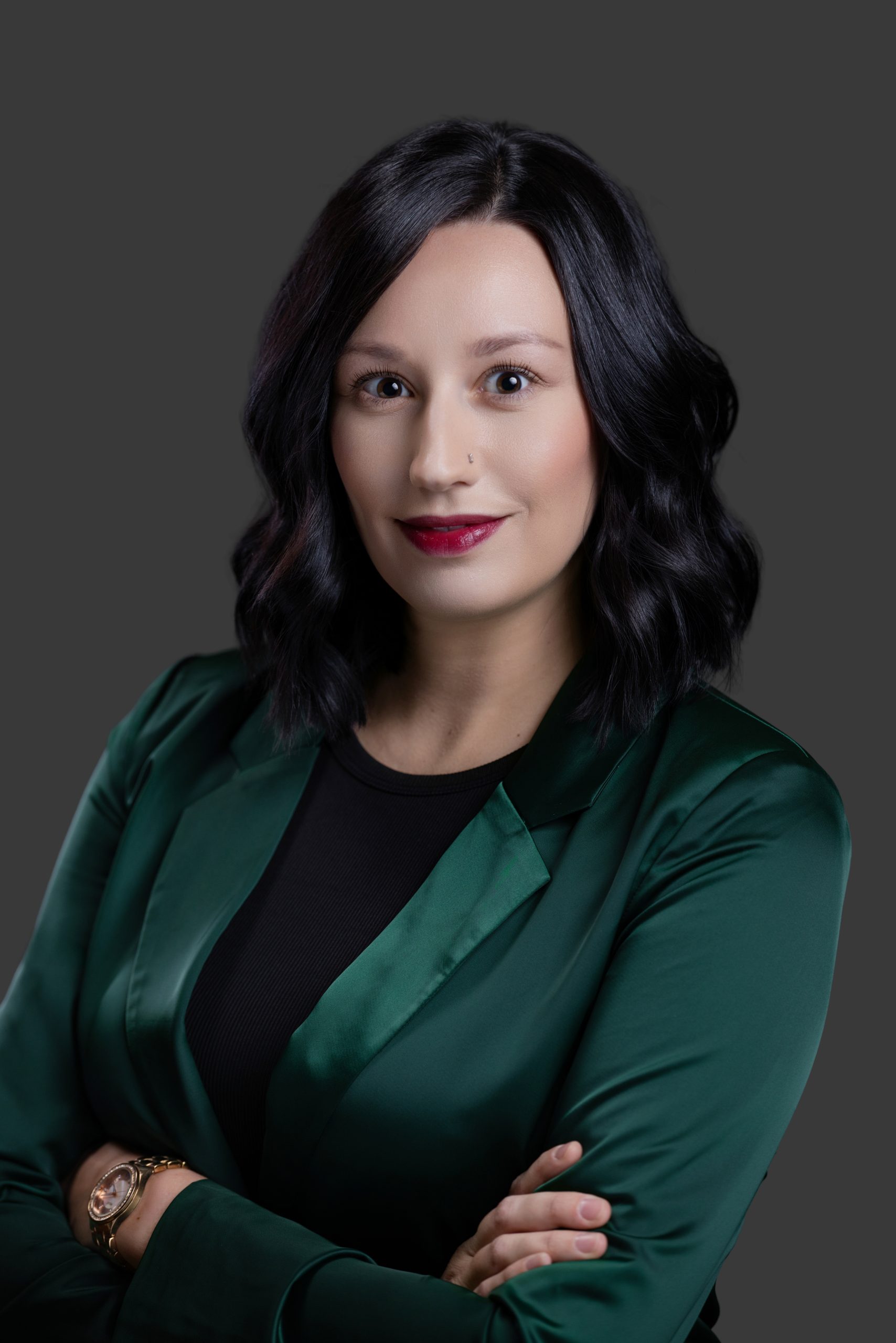 Professional studio headshot of a woman wearing a green blazer with her arms crossed against a dark gray background.