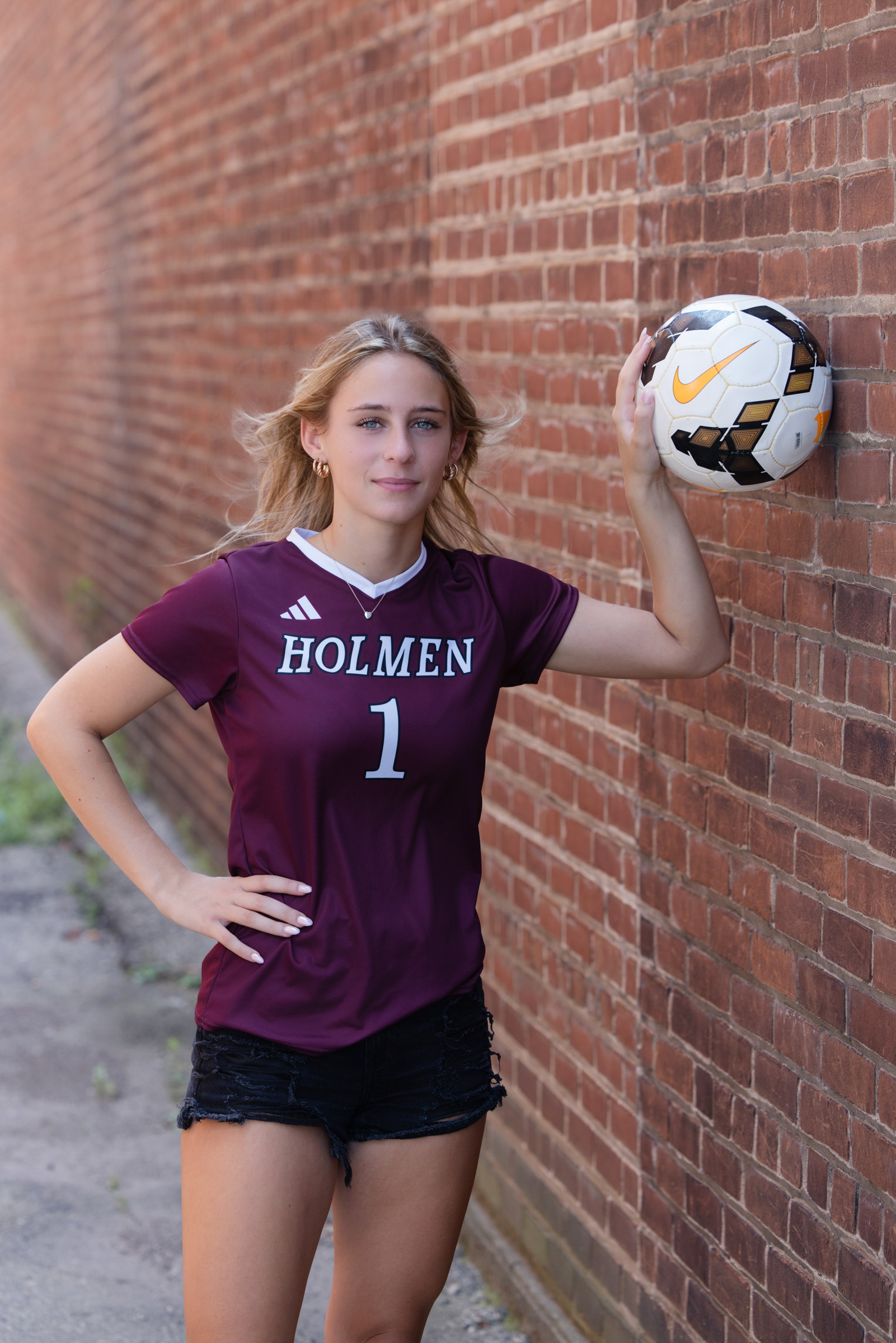 High school senior standing with a soccer ball beside a brick wall.